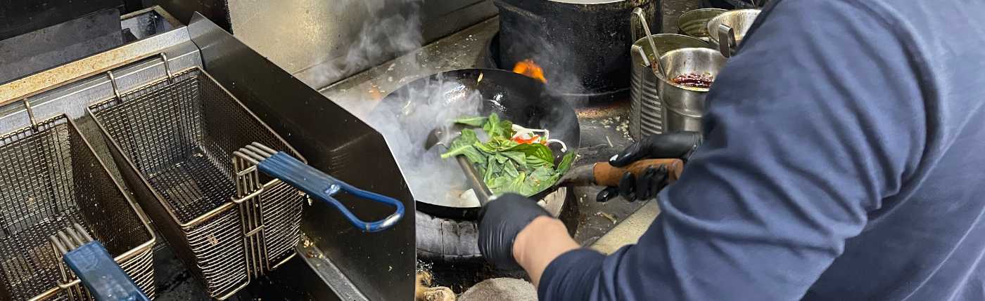 A chef in a dark shirt and gloves cooks vegetables in a steaming wok